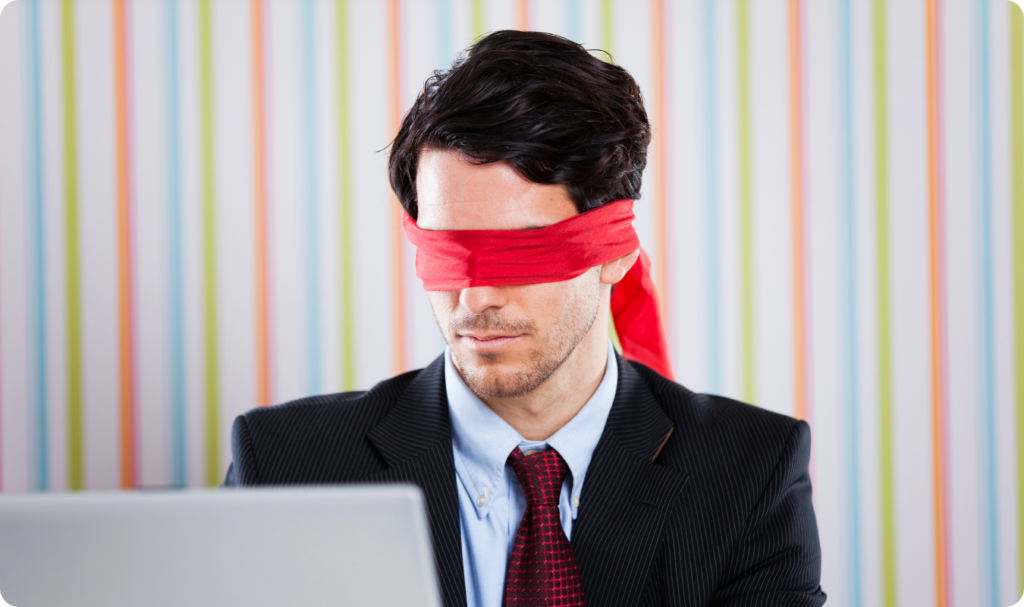 Um homem jovem, vestido com terno preto, camisa azul clara e gravata vermelha, está sentado em frente a um computador portátil (laptop). Ele está com os olhos vendados por uma faixa de tecido vermelho escarlate, simbolizando a falta de visão ou direção. O fundo é composto por um padrão de listras verticais coloridas em tons pastéis de rosa, amarelo, azul e verde. A imagem representa a metáfora de um gestor operando "no escuro", sem o auxílio de dados ou tecnologia estratégica.