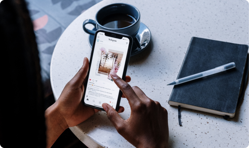 Visão de cima das mãos de uma pessoa negra segurando um smartphone e navegando pelo feed do Instagram. O celular está posicionado sobre uma mesa de mármore clara, ao lado de uma xícara de café azul, um caderno preto e uma caneta. A imagem simboliza a curadoria de conteúdo e a gestão estratégica de mídias sociais realizadas pela Vanguarda Martech para conectar marcas e consumidores