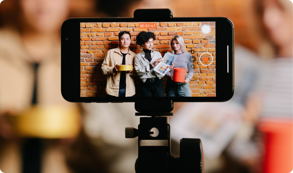 Smartphone em um tripé gravando três influenciadores em frente a uma parede de tijolos. O trio segura produtos e uma paleta de cores, enquanto a tela do celular mostra a interface de gravação ativa com cronômetro. A imagem ilustra a criação de conteúdo profissional, o uso de ferramentas de automação e a importância do vídeo nas estratégias de marketing digital modernas.
