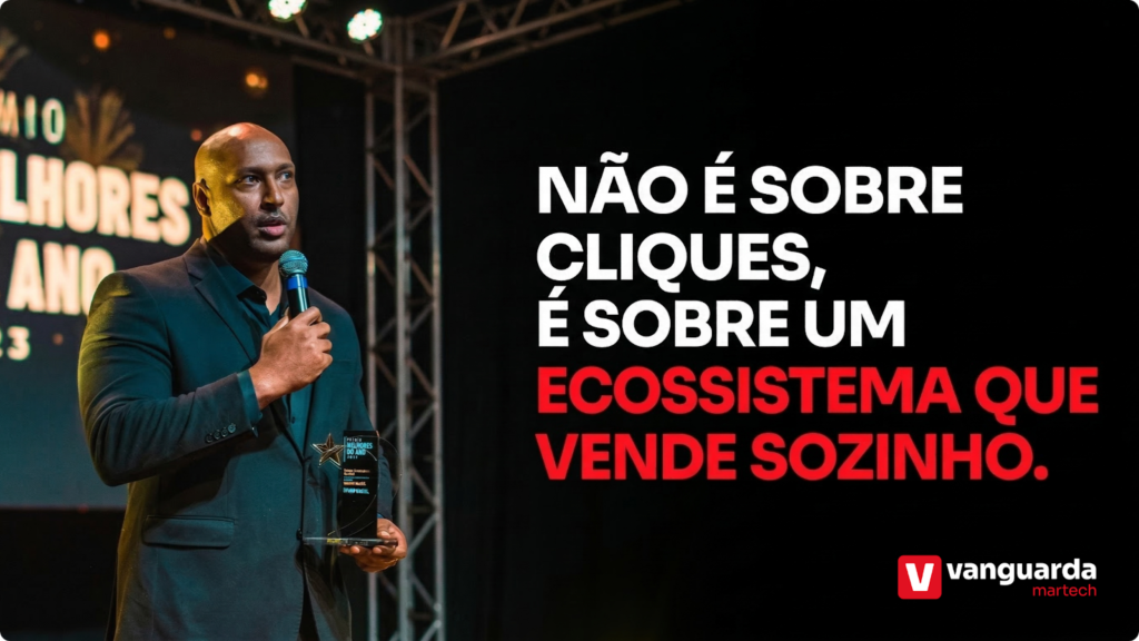Um homem negro de terno escuro segura um microfone e um troféu "Melhores do Ano" enquanto discursa em um palco iluminado. Ao lado dele, em letras grandes e de alto contraste sobre um fundo preto, lê-se a frase: "NÃO É SOBRE CLIQUES, É SOBRE UM ECOSSISTEMA QUE VENDE SOZINHO". No canto inferior direito, aparece o logotipo da Vanguarda Martech em vermelho e branco.