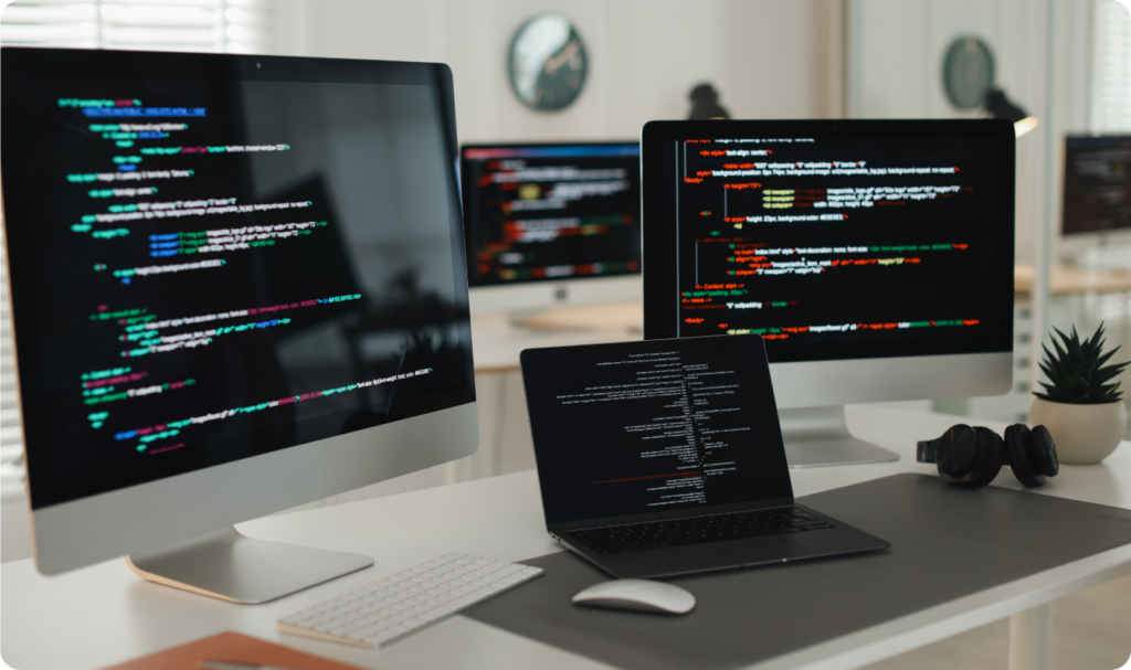 Estação de trabalho moderna em um escritório de design, apresentando um laptop aberto e dois monitores de alta resolução exibindo linhas de código de programação coloridas. O ambiente é limpo e profissional, com iluminação clara, simbolizando a excelência técnica da Vanguarda Martech em desenvolvimento web e design de interfaces.