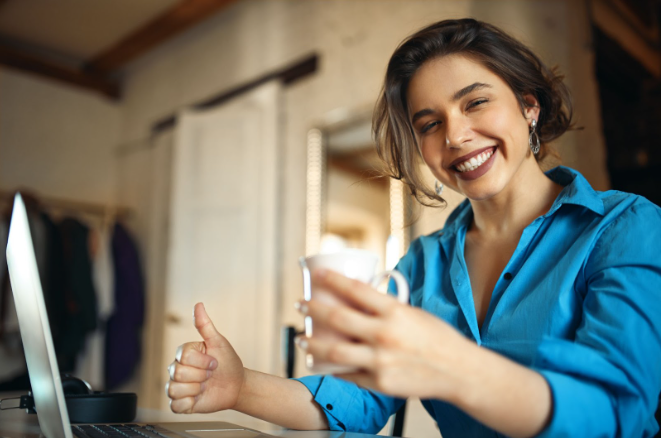 Mulher jovem e sorridente usando uma camisa azul, sentada à frente de um laptop em um ambiente de escritório moderno, segurando uma caneca e fazendo um sinal de "joinha" (thumbs up), representando o sucesso e a satisfação no atendimento ao cliente via WhatsApp.