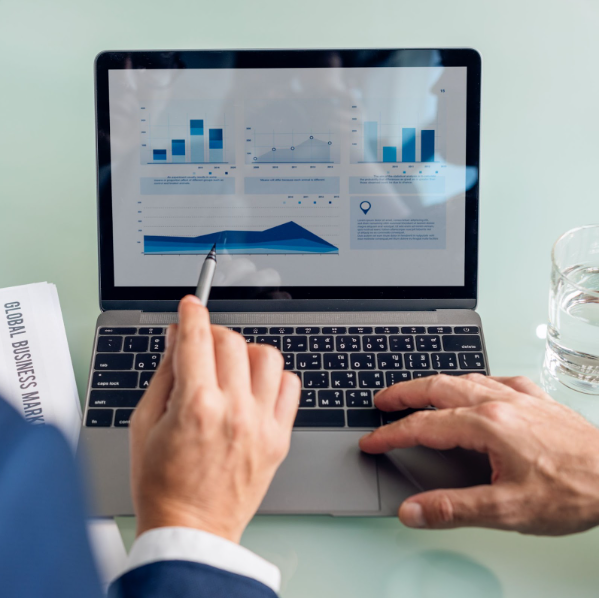 Close-up de um profissional analisando gráficos de desempenho em um laptop prata. A tela exibe diversos infográficos de barras e um gráfico de área azul crescente sob o título 'Global Business Market'. Uma mão utiliza uma caneta touch para apontar dados específicos, enquanto a outra opera o trackpad, indicando uma auditoria de métricas estratégica