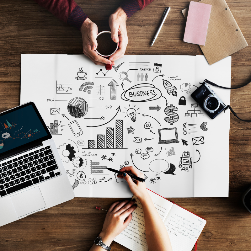 
Duas pessoas trabalhando em uma mesa de madeira. A partir de uma visão de cima, uma mão segura uma xícara de café enquanto a outra mão desenha em um grande papel com esboços de gráficos, ícones de negócios e um mapa mental. Ao lado, há um laptop, um caderno e uma câmera fotográfica, sugerindo uma sessão de brainstorming e planejamento de negócios ou marketing.